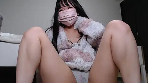 Media: Video of an Asian woman with long black hair, wearing a pink face mask, a fluffy pink jacket, and panties, seated with legs spread, in a dimly lit room with grey walls.