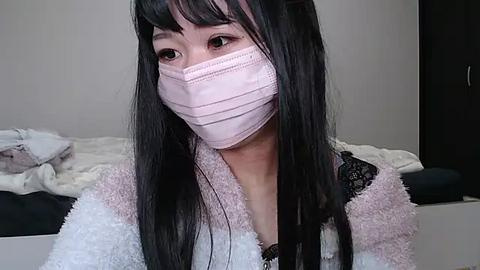 Media: Video of a young East Asian woman with long black hair, wearing a pink face mask, a white fluffy robe, and a black lace bralette, sitting in a simple room with white walls and a bed in the background.