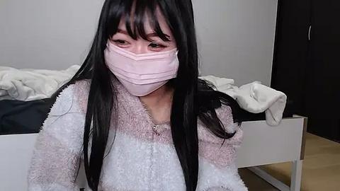 Media: Video of an East Asian woman with long black hair, wearing a pink face mask, gray sweater, and white shirt, sitting on a bed with white sheets and a dark blanket, in a minimalistic bedroom with light walls.