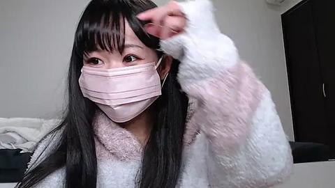 Media: Video of an East Asian woman with long black hair, wearing a pink mask and white fuzzy robe, adjusting her hair in a modern, indoor setting.