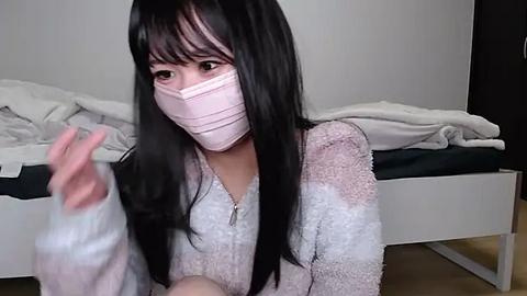 Media: Video of a young Asian woman with long black hair, wearing a pink mask, gray sweater, and a messy white bed in the background.