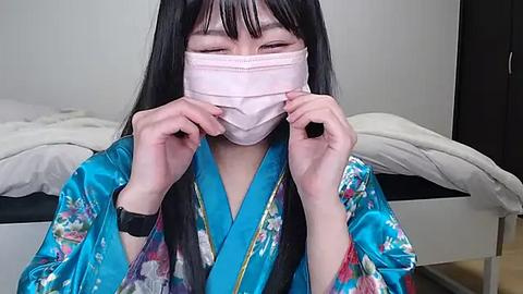 Media: Video of an Asian woman with long black hair, wearing a blue floral kimono, a pink face mask, and a black smartwatch, adjusting the mask. She is in a simple, modern bedroom with white bedding and a dark dresser in the background.