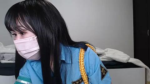 Media: Video of a young Asian woman with long black hair, wearing a blue uniform with yellow epaulets, pink mask, and white surgical gown, sitting on a bed, looking down.