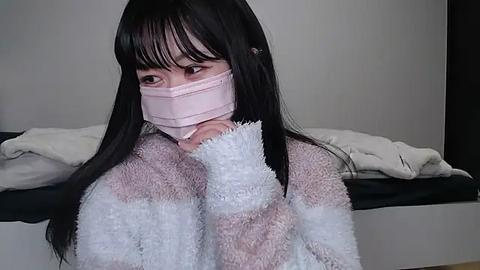 Media: Video of an Asian woman with long black hair, wearing a pink surgical mask, a white fuzzy sweater, and a black bra, sitting on a bed with white sheets, indoors.