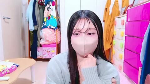 Media: Video of an Asian woman with long black hair and a beige face mask, seated in a brightly lit, organized bedroom with pink drawers and hanging clothes.