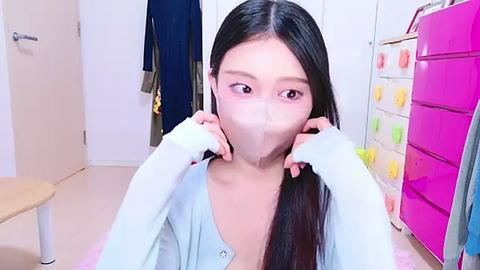 Media: A video of an Asian woman with long black hair wearing a light blue cardigan and a surgical mask, sitting in a brightly lit room with colorful storage cabinets and a wooden chair.