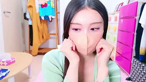 Media: Video of an Asian woman with long black hair wearing a face mask in a colorful, cluttered bedroom with a wooden wardrobe, pink drawers, and hanging clothes.