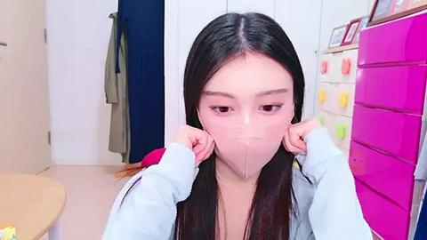 Media: Video of an Asian woman with long black hair, wearing a light blue hoodie and a face mask, adjusting her mask in a bright, organized room with white cabinets, a pink file cabinet, and a green coat hanging.