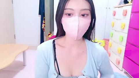 Media: Video of an East Asian woman with long black hair, wearing a pink surgical mask, light blue top, and black bra, in a brightly lit, colorful room with white walls, pink storage bins, and a wooden table.