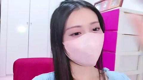 Media: Video of an Asian woman with long black hair wearing a white mask and light blue top. Background includes white cabinets and a pink storage unit.