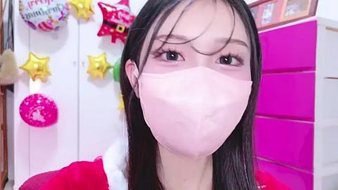 Media: Video of an Asian woman with long black hair, wearing a white face mask, red dress, and pink background with festive decorations.