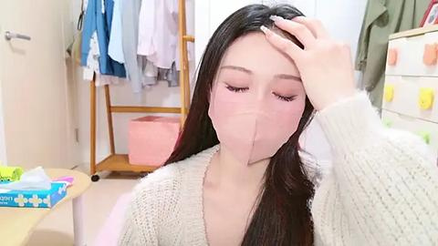 Media: Video of a young East Asian woman with long black hair, wearing a beige knit cardigan, face mask, and resting hand on her forehead in a bright, modern bathroom.