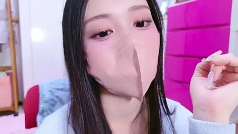 Media: Video of a young Asian woman with long black hair, wearing a light blue sweater, holding a large piece of paper over her face, in a bright pink room with a red chair.