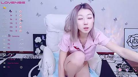 Media: A video of an Asian woman with light skin, shoulder-length blonde hair, and closed eyes, wearing a pink shirt, sitting on a bed with floral-patterned bedding, with a \"LoveSense\" streaming platform in the background.