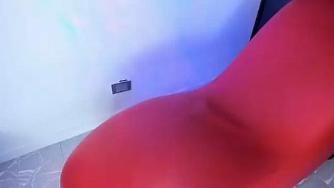 Media: Video of a modern, red, curved chair in a minimalist room with a white wall and blue light. The chair has a smooth, matte texture and is placed on a light gray tiled floor.