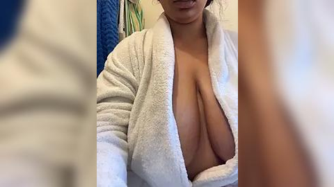 Media: A video of a woman with a medium-brown complexion, wearing a white bathrobe open to reveal her large, natural breasts. The background shows a blurred bathroom scene with blue and green towels.