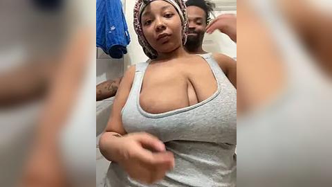 Media: Video of a curvy woman with large breasts, wearing a low-cut grey tank top, and a headscarf, standing in a bathroom.