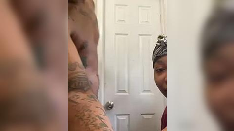 Media: A video of a tattooed, shirtless man with light skin, partially obscured by a blurred figure, standing in a bathroom with a white door and a red towel.