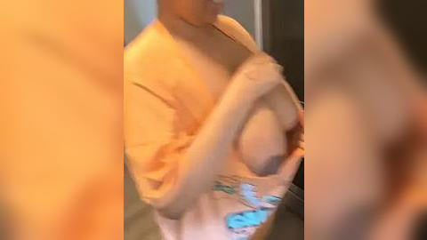 Media: Video of a light-skinned woman with medium-sized breasts, wearing a white robe, partially undressing. Her face is obscured by the robe. Background is out of focus, suggesting an indoor setting.