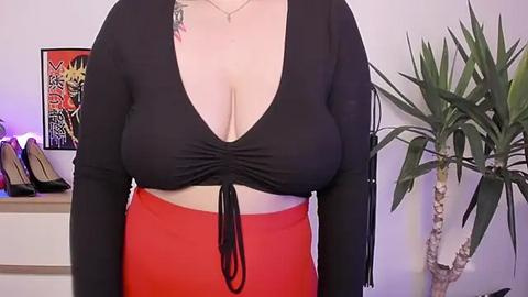 Media: Video of a curvy woman with fair skin, wearing a black halter top and high-waisted red pants, with a colorful tattoo on her left shoulder. Background features a potted plant and colorful artwork.