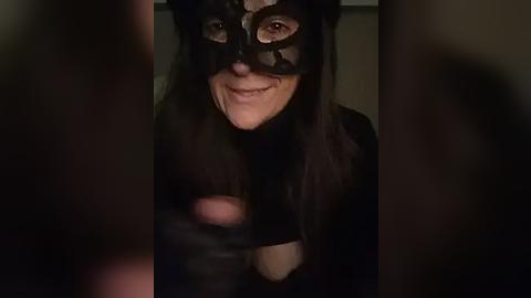 Media: Video of a woman wearing a black mask and long, dark hair, smiling seductively, with blurred, out-of-focus background.