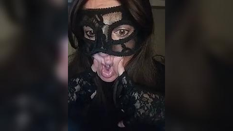 Media: Video of a woman in a black lace mask with a wide-open mouth, revealing pink tongue and teeth. She has long brown hair, and wears a black lace dress.