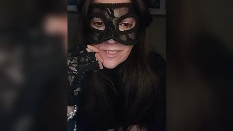 Media: Video of a woman with long brown hair, wearing a black lace mask and black gloves, smiling playfully. Background is dark, blurred.