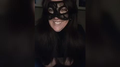 Media: Video of a smiling person wearing a black lace mask, with long dark hair, in dimly lit room.