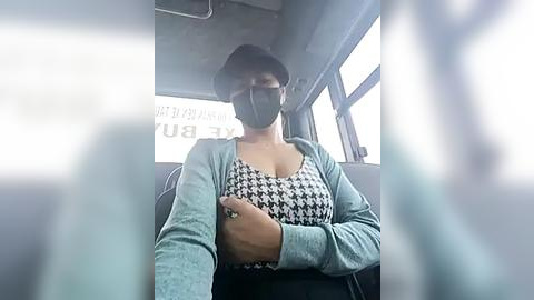 Media: Video of a woman wearing a black face mask, black and white houndstooth-patterned top, and light blue cardigan, seated in a bus with blurred passengers in the background.