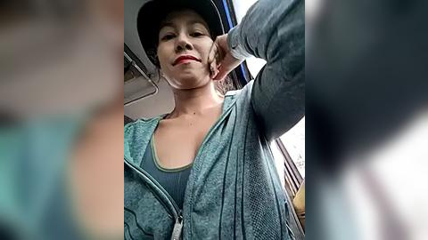 Media: Video of a young woman with light skin and dark hair, wearing a teal hoodie, black hat, and red lipstick, standing in a train with blurred passengers in the background.