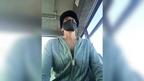 Media: Video of a person in a green hoodie, wearing a black face mask, sitting inside a bus. The image is slightly blurred, focusing on the person's face.