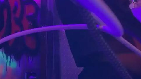 Media: A dimly-lit video of a person with long, wavy hair and a dark complexion, wearing a black shirt, standing in front of a colorful, abstract wall. The image is blurred and abstract, with purple and blue hues dominating.