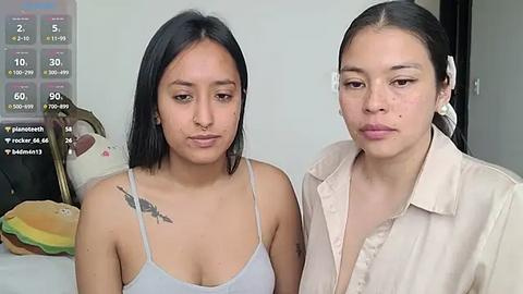 Media: Video of two young Asian women with medium skin tone, one with a tattoo on her shoulder, wearing casual outfits, in a minimalistic room.