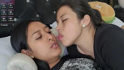 Media: Video of two women, one Asian with long black hair and a nose ring, the other Caucasian with dark hair, kissing on a bed with a tufted headboard.