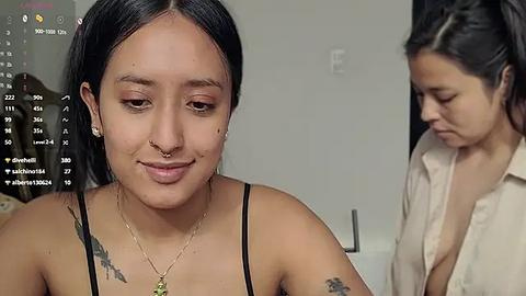 Media: Video of a young woman with light brown skin and long black hair, wearing a black bra and a necklace, sitting in a dimly lit room with a second woman partially visible in the background.