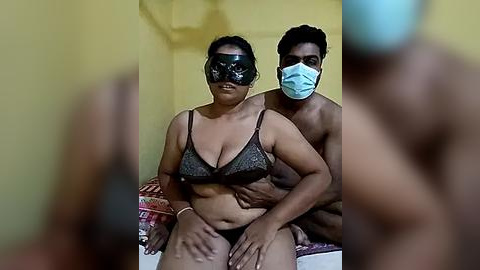 Media: Video of a muscular Black man wearing a mask and a maskless Black woman in a black lace bra, sitting on a bed in a yellow room.