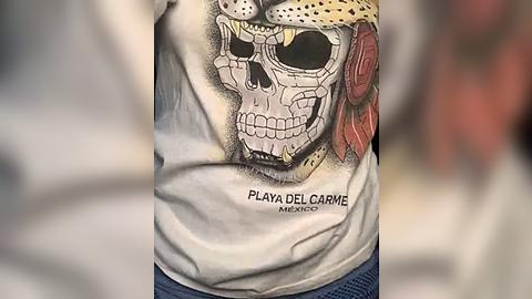 Media: Video of a person wearing a white t-shirt featuring a detailed, colorful skull design with a red flower and the text \"PLAYA DEL CARMEN MEXICO\" in black. The background is blurred, focusing on the t-shirt.