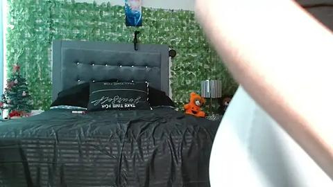 Media: A video of a modern bedroom with a gray upholstered headboard, green wall, black bedding, a stuffed animal, and a Christmas tree in the background.