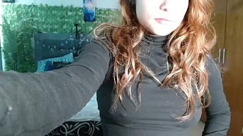 Media: Video of a fair-skinned woman with long, wavy red hair, wearing a black turtleneck, taking a selfie in a bedroom with green wall art, a wooden dresser, and a bed.