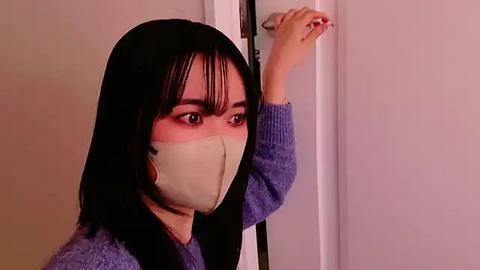 Media: Video of an Asian woman with straight black hair, wearing a beige mask and a blue sweater, leaning against a white door, with a worried expression.