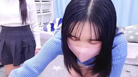 Media: Video of a young Asian woman with long black hair, wearing a blue ribbed sweater, gagged with a white cloth, kneeling on a grey carpet in a bedroom with light blue curtains and a white shelving unit.