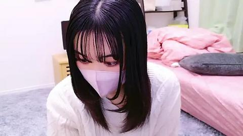 Media: A video of an Asian woman with straight black hair, wearing a white mask, a white sweater, and a white top, kneeling on a gray carpet in a pink bedroom.