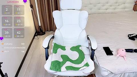 Media: Video of a white gaming chair with a green pillow, positioned in a modern bedroom with beige curtains and a white tufted headboard. A pink stuffed animal and black phone are on the bed.