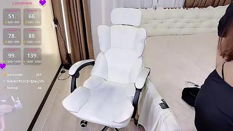 Media: Video of a modern, white ergonomic office chair in a sleek, minimalist room with beige walls and a white tufted headboard. The chair has adjustable armrests and a high backrest. A woman with brown hair, wearing a black top, sits on the right.