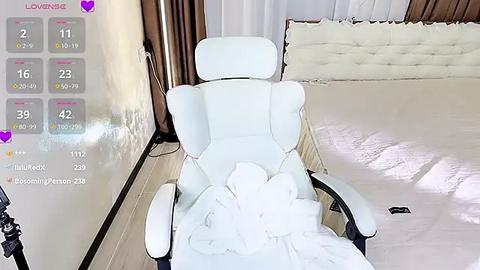 Media: A video of a white gaming chair with a plush white blanket, positioned in a modern, minimalist bedroom with beige walls and a tufted white headboard.