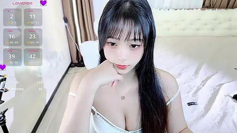 Media: Video of an East Asian woman with long black hair, wearing a white spaghetti-strap top, in a bedroom with a white bed and brown curtains.