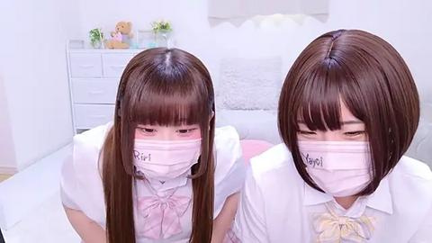 Media: Video of two young East Asian girls with straight brown hair, wearing white school uniforms, pink face masks, and name tags. They sit on a bed in a white room with a dresser and stuffed animals in the background.