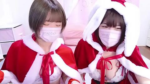 Media: Video of two Asian women with short brown hair, wearing Santa hats, red dresses, white face masks, and white lingerie, seated indoors, smiling.