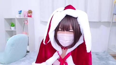 Media: Video of a young woman with short brown hair, wearing a red and white Santa hat, a white mask, and a pink dress, in a bright, white room with a blue chair and shelves.