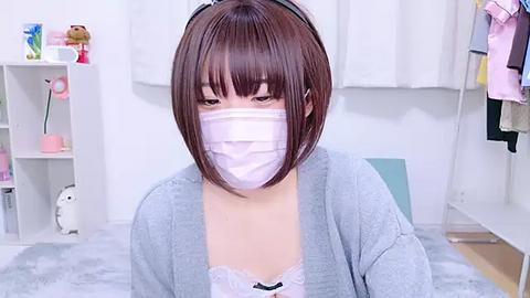 Media: Video of an East Asian woman with short brown hair, wearing a white surgical mask, gray cardigan, and lace bra, seated in a modern, minimalistic bedroom with white walls and shelves.